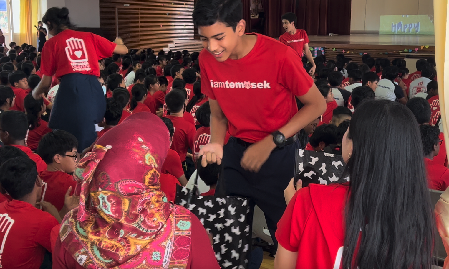 A smiling teen in a red iamtemasek shirt leans toward seated students in a crowded school hall event.